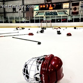 An indoor ice hockey rink with various pieces of hockey equipment scattered on the ice, including helmets and sticks. Banners and flags adorn the walls above the rink, and a scoreboard displays the end of a game.