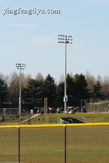 A sports field with a group of people engaging in athletic activities in the background. There is sports equipment like a soccer goal and a scoreboard visible. A chain-link fence runs across the foreground, and a statue or sculpture resembling an eagle is also seen. Trees and tall floodlights are positioned behind the field.