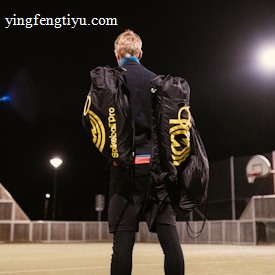A person stands on an outdoor sports court at night, carrying two large duffle bags labeled 'Spikeball Pro.' The court is lit by bright overhead lights and features a basketball hoop and a fence.