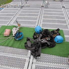 Climbing gear including helmets, harnesses, ropes, and other equipment is arranged on a green mat placed over a metal grid surface. The scene includes climbing helmets in blue and various metal carabiners and tools. A person's leg is visible on the right side, suggesting preparation or presence of a person for an upcoming activity.
